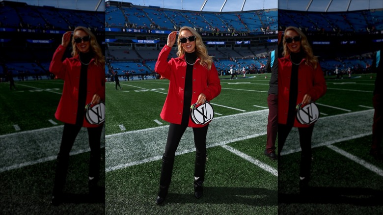 Brittany Mahomes, posing with her hand up, standing on the sidelines of a football field.