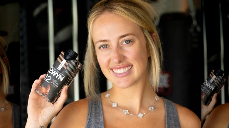 Brittany Mahomes in a gray workout set, smiling and holding a protein drink.