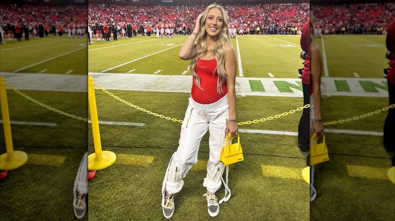 Brittany Mahomes poses on the field in a red tank top and white pants