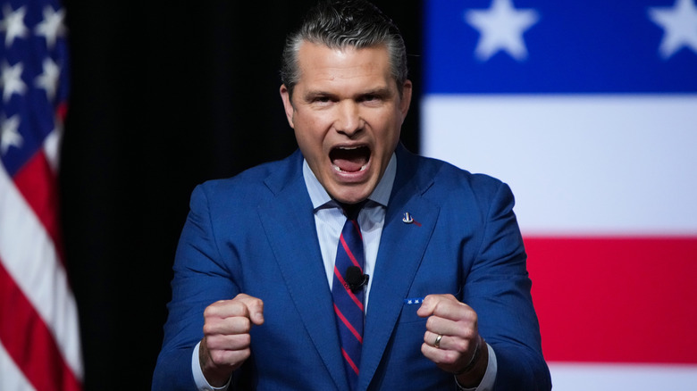 Pete Hegseth yelling with clenched fists in a navy suit in front of an American flag