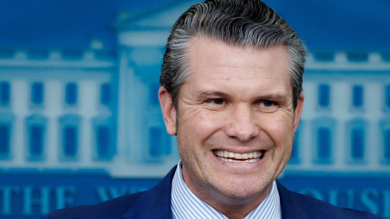 Pete Hegseth forcing a smile in a navy suit with a stripy blue and white shirt