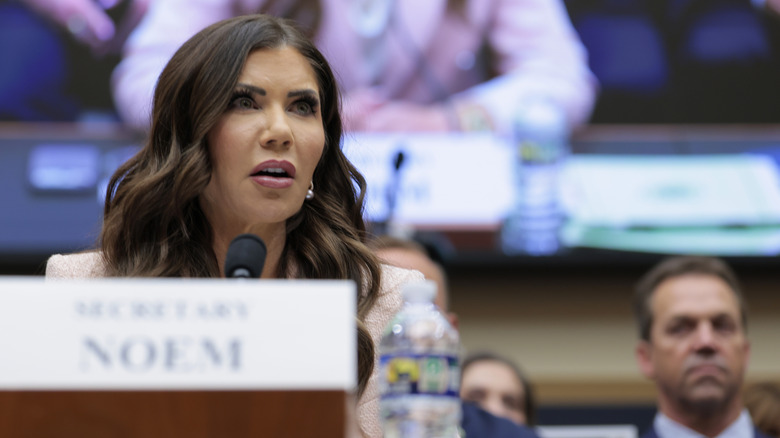 Kristi Noem addressing the judges during her hearing while Bryon Noem watches out of focus behind her