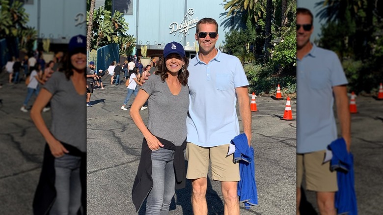 Bryon Noem smiling while wearing sunglasses, a light blue shirt, and tan pants while next to Kristi Noem who's wearing a gray t-shirt, blue pants and a cap.