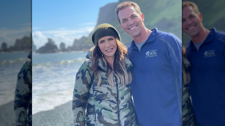 Kristi Noem smiling in a green jacket and hat next to Bryon Noem who's wearing a blue sweater and shirt.