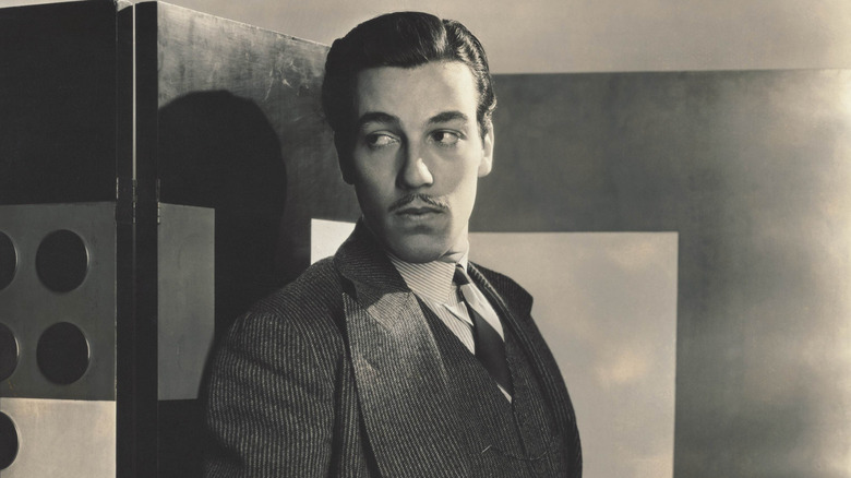 Cesar Romero, with a thin mustache and dark hair, wearing a dark striped three-piece suit, with a striped shirt, and a striped tie, standing with his back against a wall, with both hands in his pockets, looking to the side.