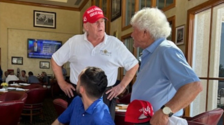 Donald Trump in a red MAGA hat speaking to two guests at Mar-a-Lago