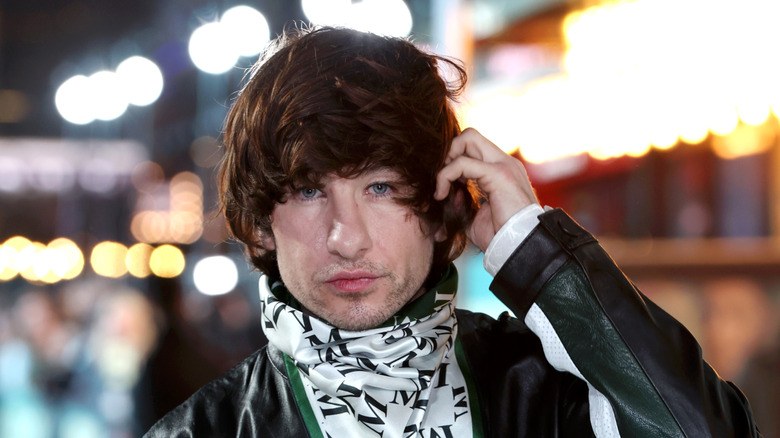Barry Keoghan holding his hand to his moppy brown hair, pursing his lips, and wearing a silk M monogrammed scarf over a black and green motorcycle jacket
