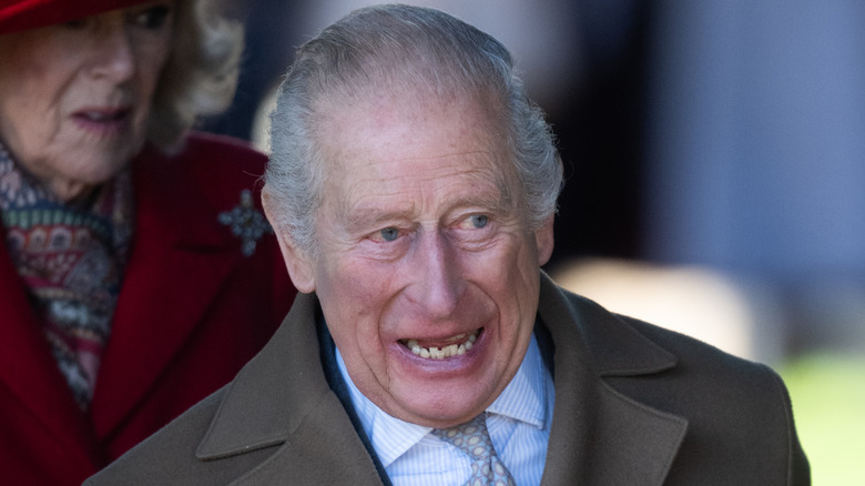 Closeup of King Charles III laughing while Queen Camilla looks concerned in background