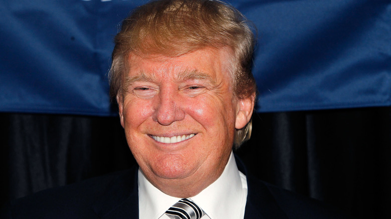Donald Trump wearing a suit and striped tie and smiling in front of a dark background with a blue curtain
