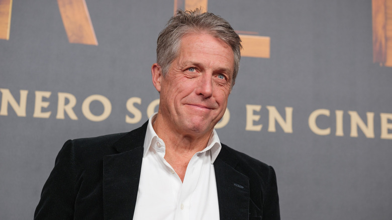 Hugh Grant smiling and wearing a black blazer over a white shirt