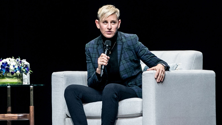 Ellen DeGeneres speaking onstage at an event in a pinstripe suit