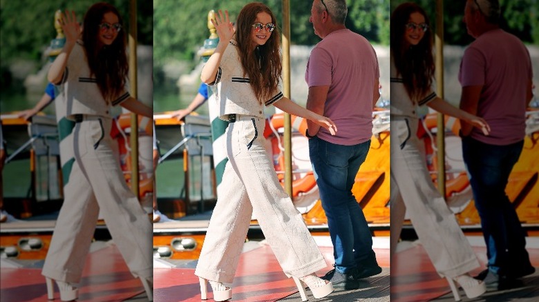Sadie Sink in white outfit and sunglasses waving to camera