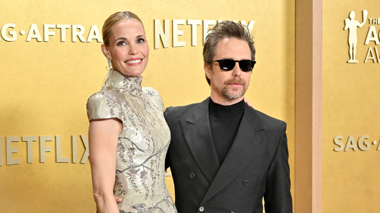 Sam Rockwell in sunglasses and black tux beside Leslie Bibb