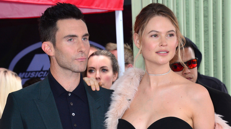 Adam Levine unsmiling in dark suit and Behati Prinsloo in strapless black dress and pink fur coat