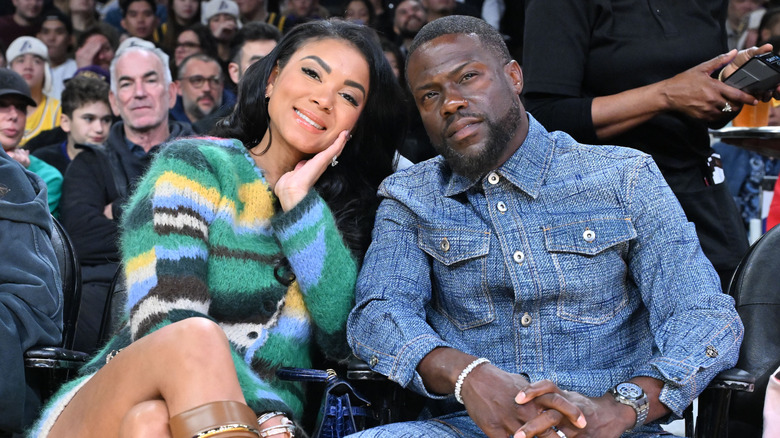 Eniko Hart in striped sweater sitting next to Kevin Hart in blue shirt at a basketball game