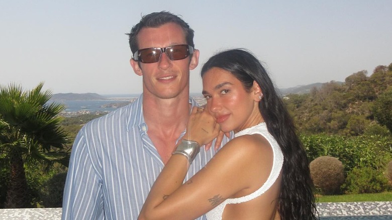 Callum Turner in sunglasses with Dua Lipa leaning on his shoulder, both smiling.