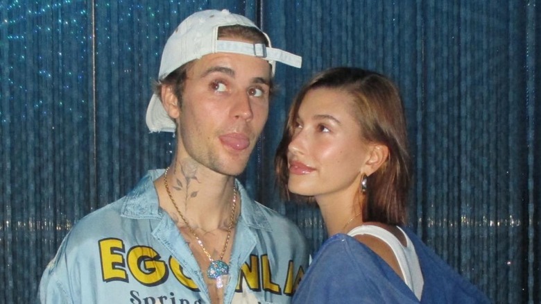 A close-up of Justin Beiber sticking out his tongue with Hailey Bieber looking on.