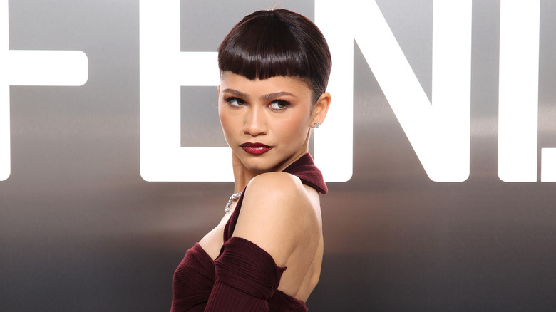 Zendaya with a fringe hairstyle wearing a maroon dress looking over her shoulder