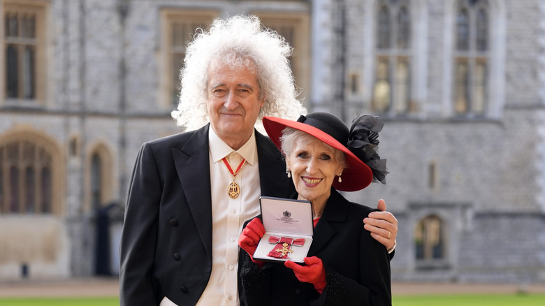 Brian May with his arm around wife Anita Dobson, holding the knighthood medal