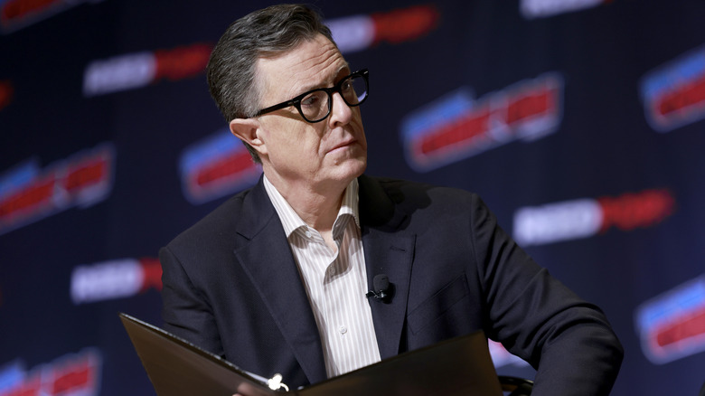Stephen Colbert with a bent arm and an open leather folder, looks off to the right.