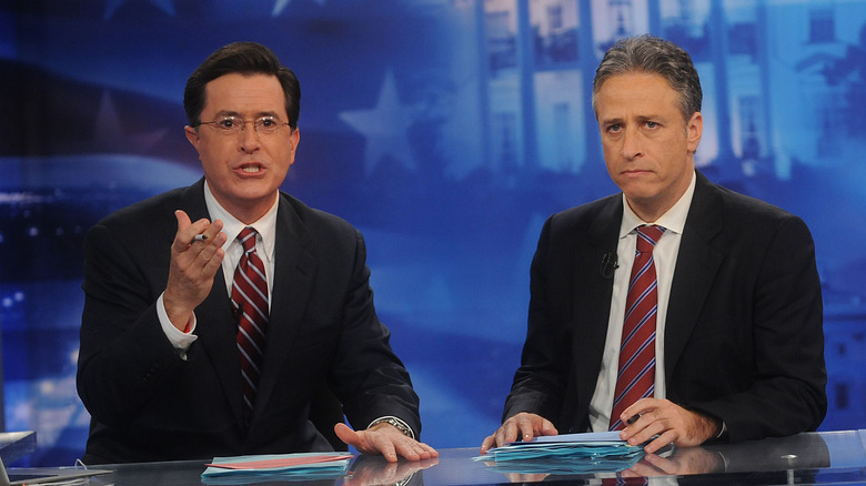 Stephen Colbert and Jon Stewart sit at a desk