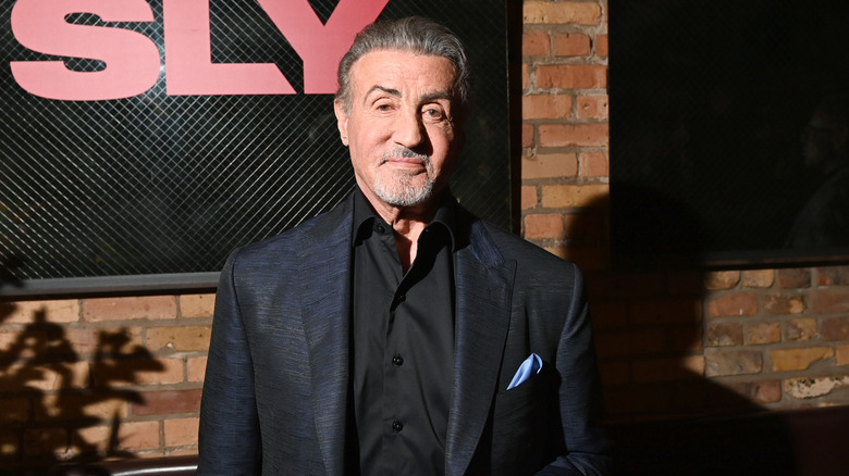 Sylvester Stallone in a blue formal blazer against an exposed brick background