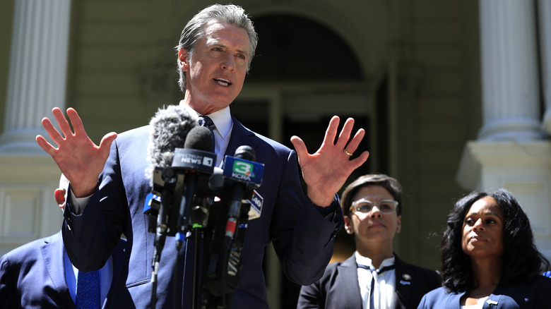 Gavin Newsom speaking to reporters in a suit while raising his hands in the air