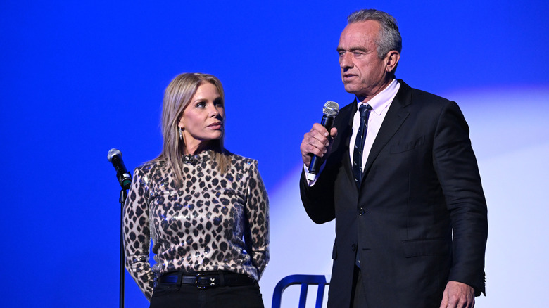 Cheryl Hines looking at RFK Jr. speaking into a microphone in a suit