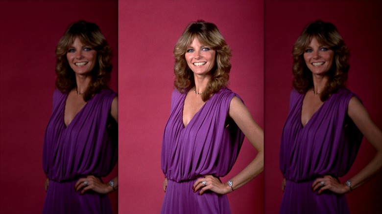 Cheryl Tiegs posing with her hands on her hips in a purple dress against a burgundy background, with her mousey brown hair curled with curtain bangs