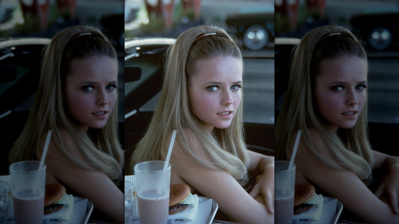 Cheryl Tiegs with long, straight hair pulled back in a headband posing in a drop-top car with a burger and milkshake beside her