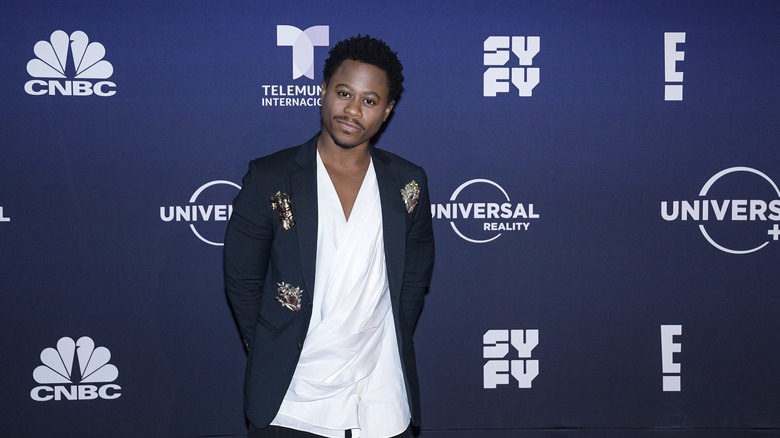 Daniel Kyri posing in front of a blue NBC network background wearing a black jacket and white silk shirt.
