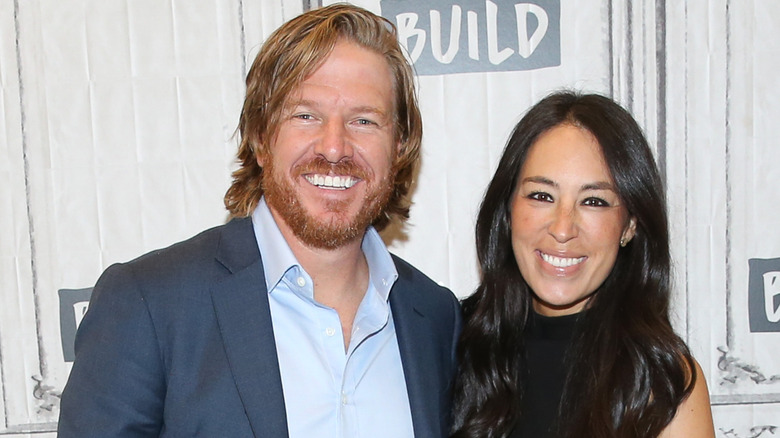 Chip Gaines with long hair posing with his wife Joanna Gaines in front of a white background.