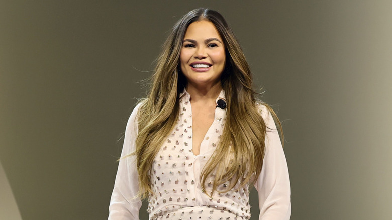 Chrissy Teigen smiling onstage in a white and gold outfit