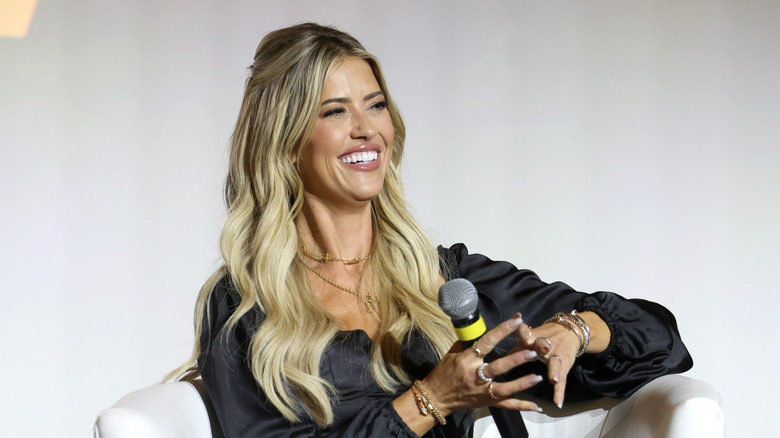 Christina Haack smiling while sitting down and holding a microphone in a black shirt