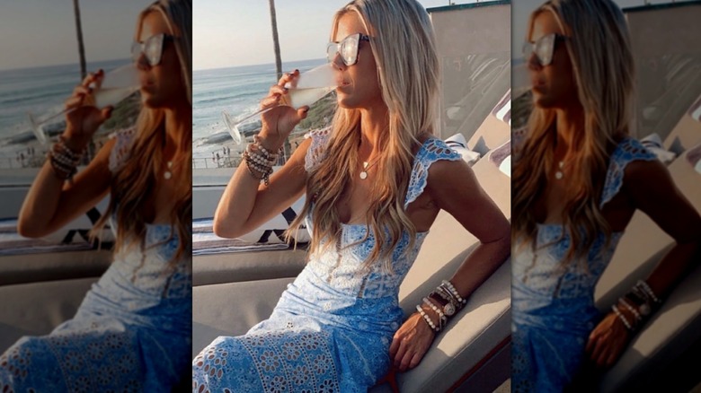 Christina Haack in a blue dress wearing sunglasses while drinking champagne on a sun lounger in front of a sea view