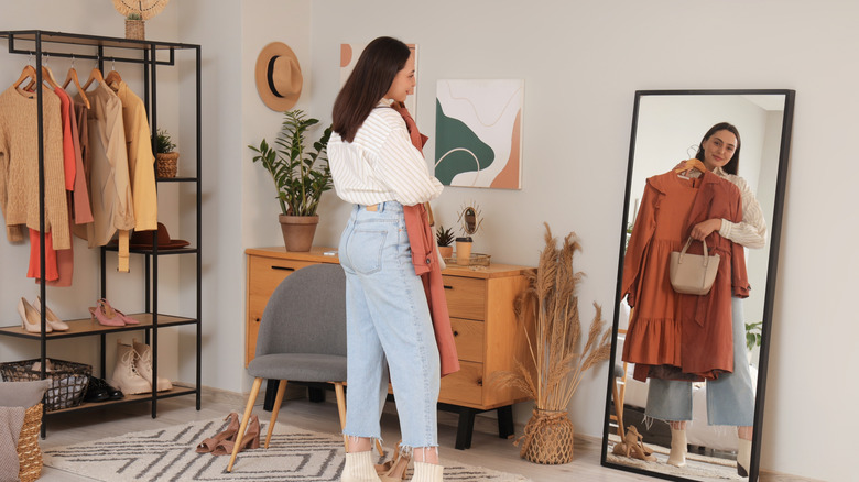 Young woman trying on stylish clothes with bag near mirror at home