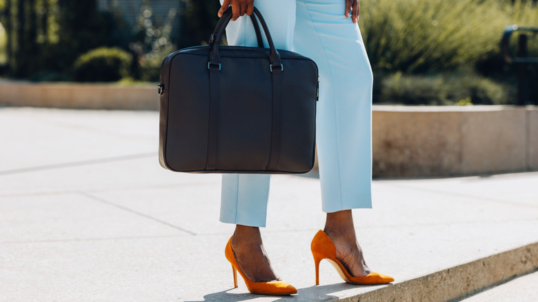 Lower half of woman standing outside wearing blue suit, orange heels, and holding black briefcase