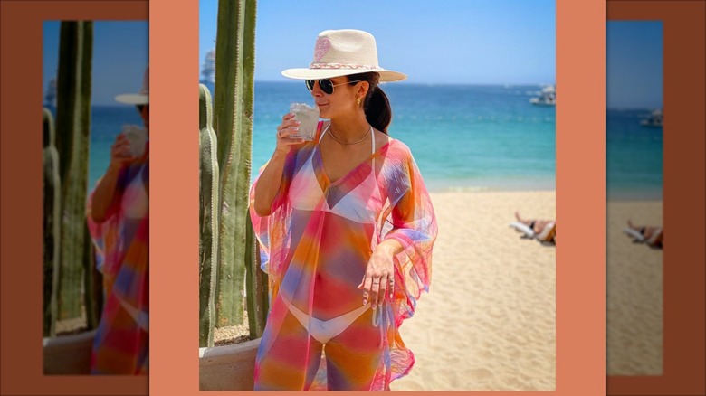 Kaitlan Collins sipping out of a glass while wearing dark sunglasses, a white cowboy hat, and a white bikini under a transparent gown.