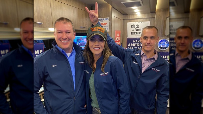 Eric Held,and Kristi Noem smiling together while Corey Lewandowski making a devil horn gesture above her head