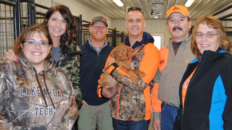 Kristi Noem smiling and Corey Lewandowski smiling while holding a dog with a group of people