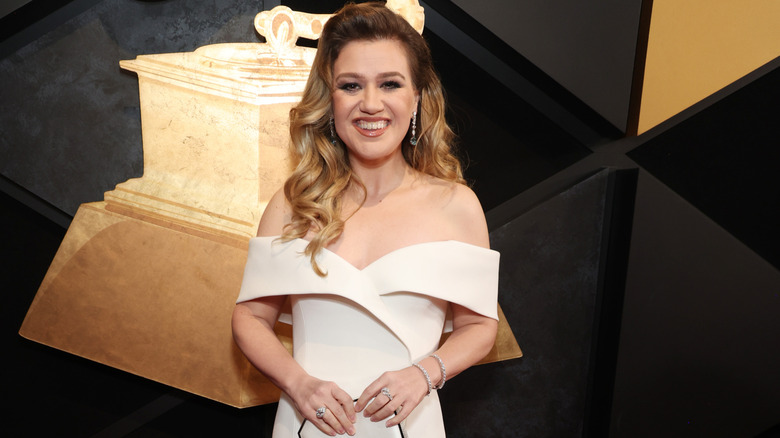Kelly Clarkson smiling as she poses on the red carpet in a white off the shoulder dress