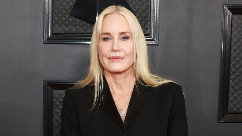 Daryl Hannah stands on the red carpet against a black background with her lips closed and blonde hair down wearing a black shirt with a peace sign necklace