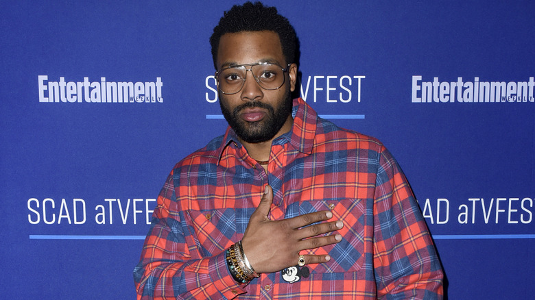LaRoyce Hawkins wearing a plaid shirt and posing with his hand on his chest.