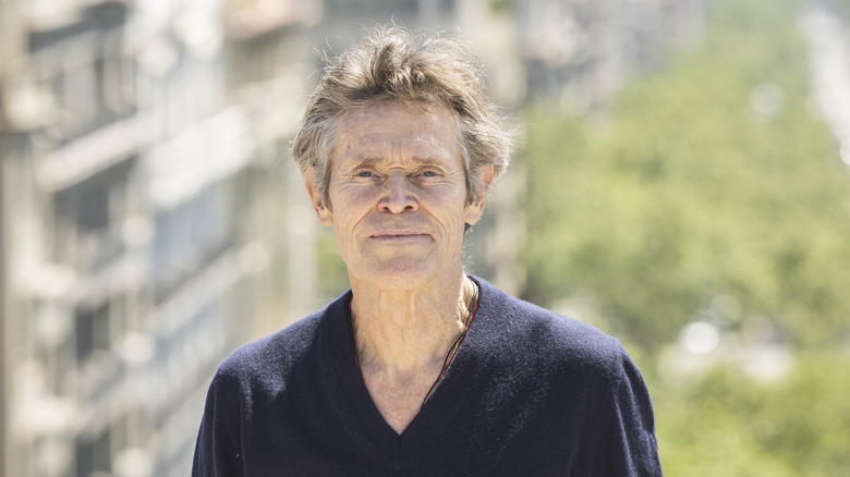 Willem Dafoe smiling in a plain navy V-neck sweater
