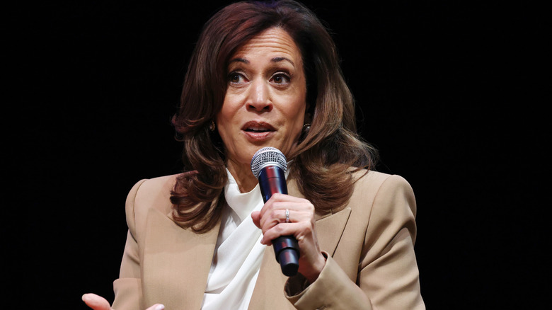 Kamala Harris speaking into a microphone in a tan jacket with a white shirt underneath