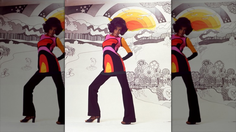 Diana Ross posing for a photo shoot in black flares and a colorful long sleeved mini dress against an illustrated background
