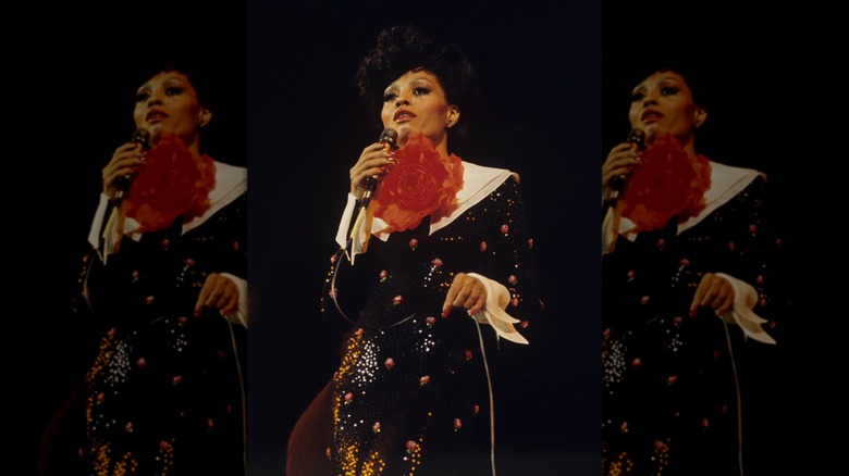 Diana Ross performing in an embroidered dress with a wide white color and massive rose blooming from her neck