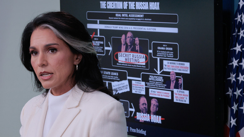Tulsi Gabbard addressing the media in a white suit with a visual of the so-called "Russia Hoax" depicted behind her