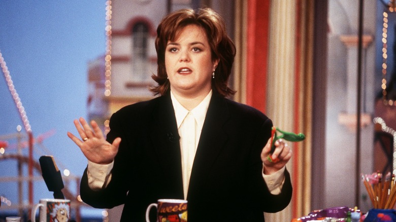 Rosie O'Donnell in a suit, sitting behind the desk on her talk show.