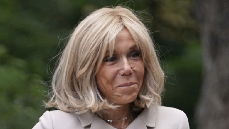 Brigitte Macron smirking while wearing a white dress and skirt at Downing Street in London.